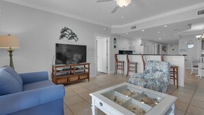 A living room with a television, a blue sofa, a coffee table with decorative items, a ceiling fan, and a kitchen area visible through an open door.