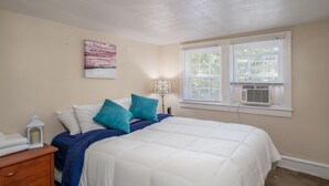 A bedroom with a bed, a nightstand, a lamp, a window with blinds, and a wall-mounted air conditioner.