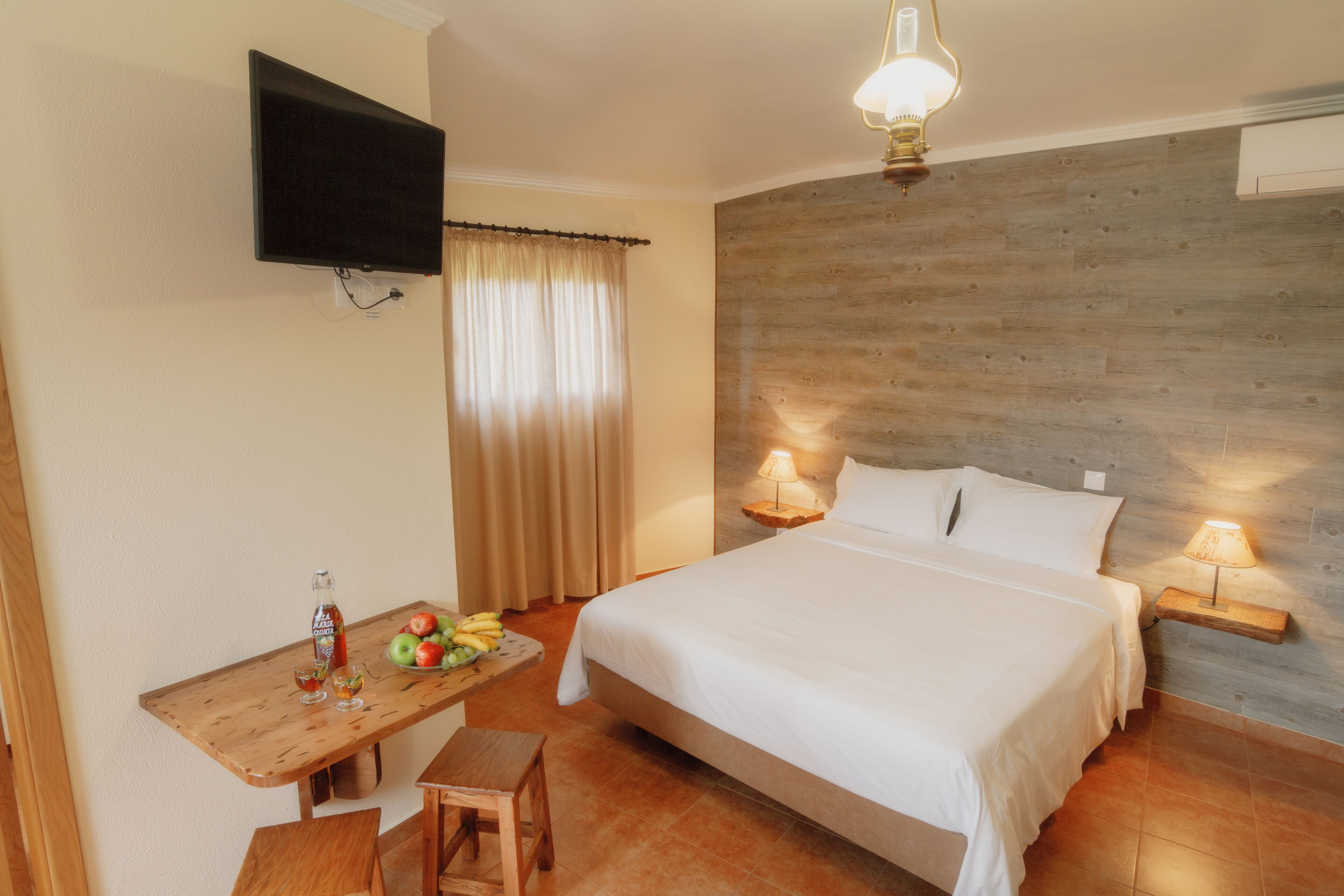 Habitación de hotel con una cama, lámparas de noche, un televisor montado en la pared, un escritorio de madera con una botella y fruta, y una ventana con cortinas.