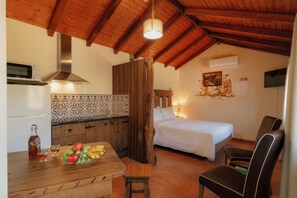 A cozy room with a bed, a wooden table with fruit, a chair, and a kitchenette.