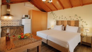 Une pièce avec un plafond en bois, un lit avec une literie blanche, un coin cuisine équipé d’un micro-ondes, et une table avec des fruits et une bouteille.