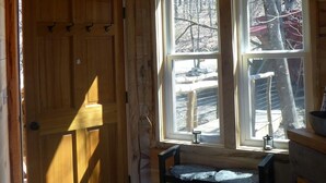 Interior de una cabaña de madera con una puerta grande de madera, dos ventanas que ofrecen vistas a un paisaje nevado, una cesta tejida colgando del techo y un banco junto a las ventanas.