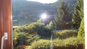 Vista a través de una ventana con una puerta, mostrando un paisaje con colinas, árboles y un sol brillante.