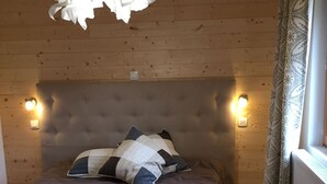 Un dormitorio con cabecera capitonada, colcha gris, almohadas estampadas, lámpara colgante y dos luces fijadas a la pared.