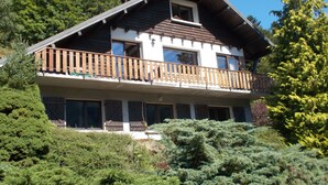 Un chalet de dos pisos con balcón, rodeado de abundante vegetación.