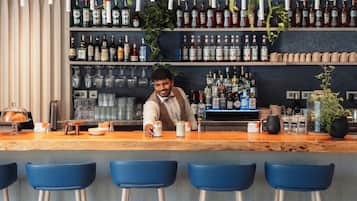 A bar area with a bartender, shelves of liquor bottles, hanging plants, and blue bar stools.