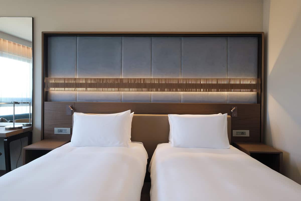 Habitación de hotel con dos camas, cabecero de madera y mesita de noche con lámpara.