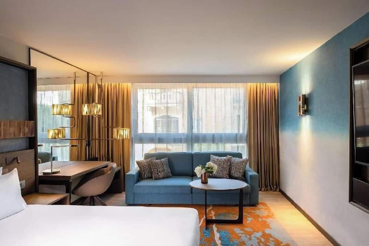Habitación de hotel con cama, sofá, escritorio y una mesita. Hay cortinas en las ventanas y una lámpara sobre el escritorio.