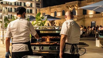 Dos cocineros preparando comida al aire libre de noche, con un edificio e invitados al fondo.