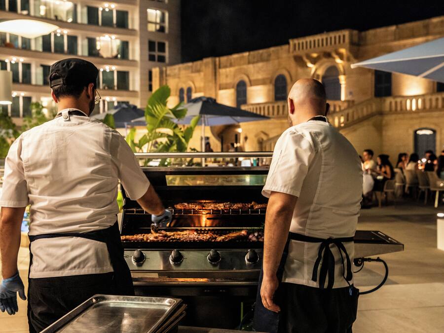 Dos cocineros preparando comida al aire libre de noche, con un edificio e invitados al fondo.