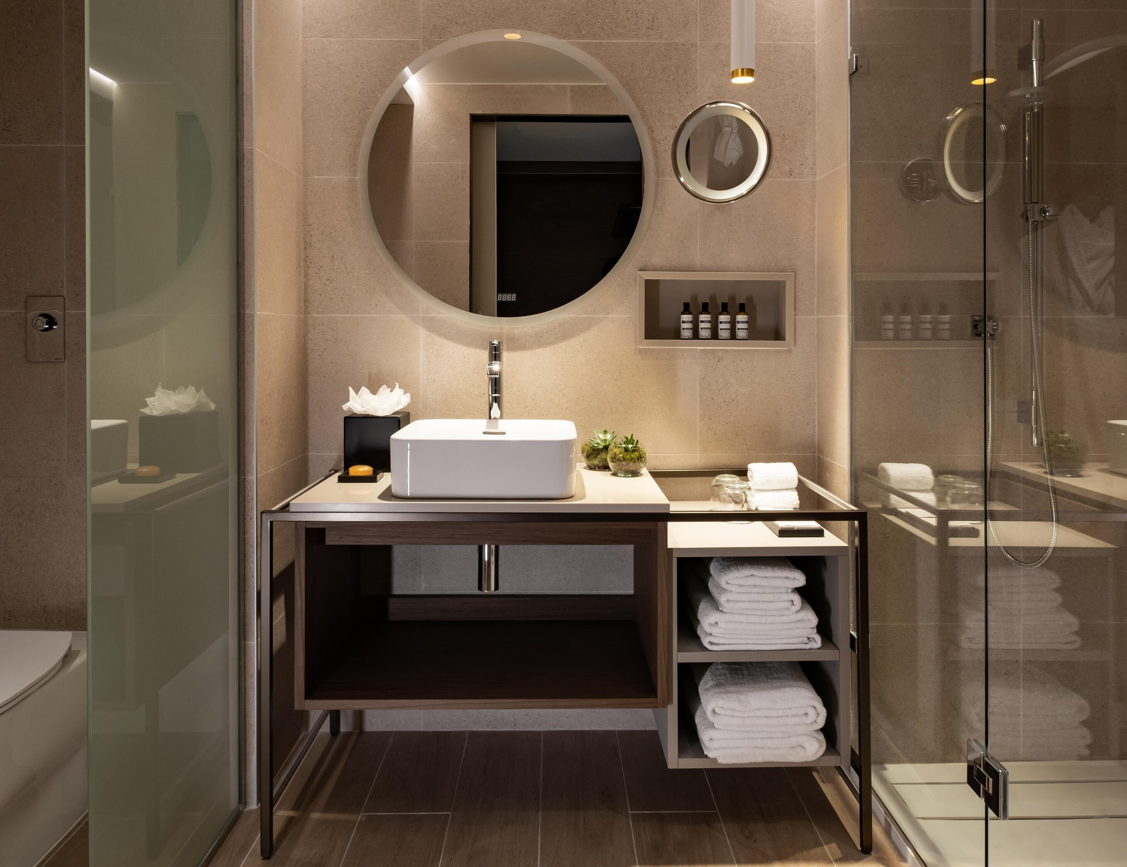 A modern bathroom with a glass-enclosed shower, a round mirror, a white sink, and neatly folded towels.