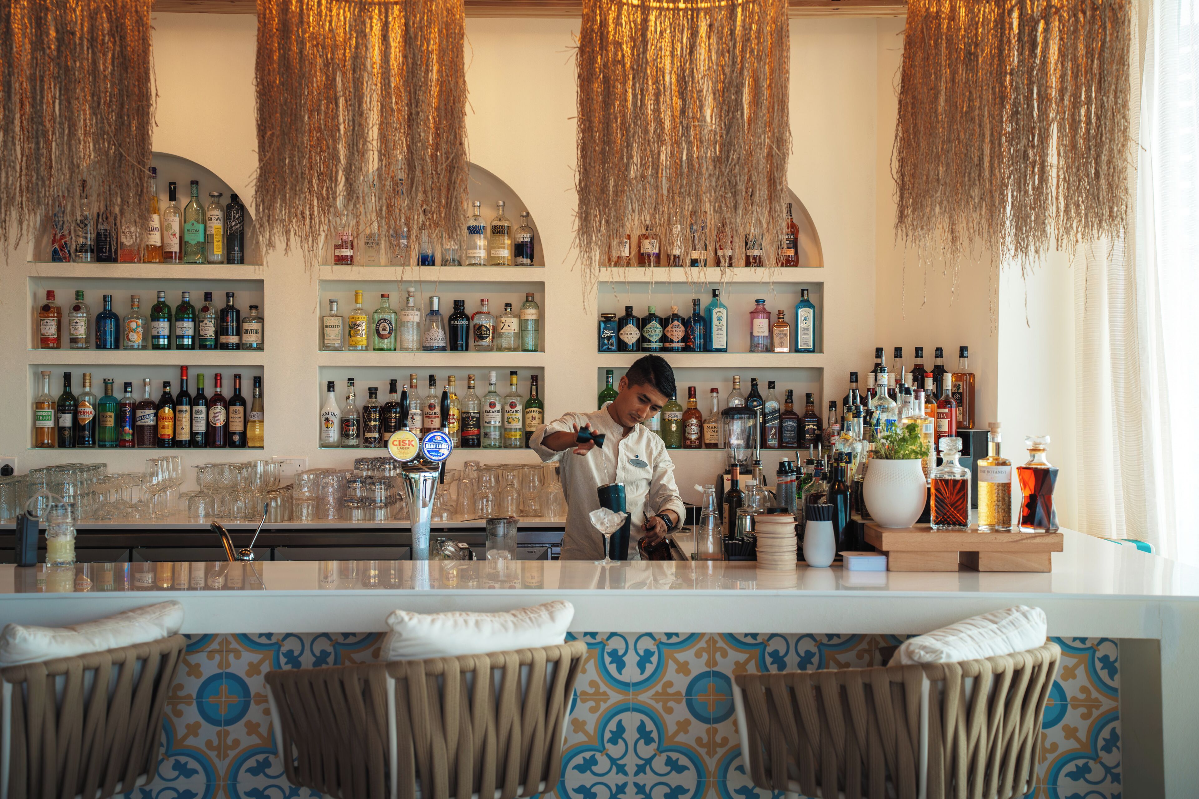 A bar with a bartender preparing drinks, shelves of liquor bottles, and a patterned wall.