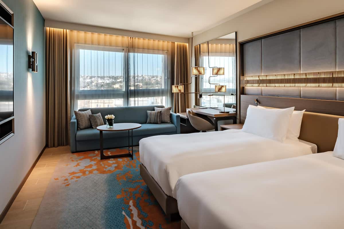 Habitación de hotel con dos camas, un sofá, un escritorio y vista a la ciudad.