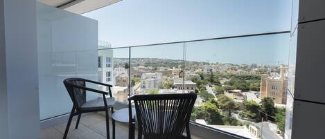 A balcony with two chairs and a small table, overlooking a cityscape with buildings and greenery.