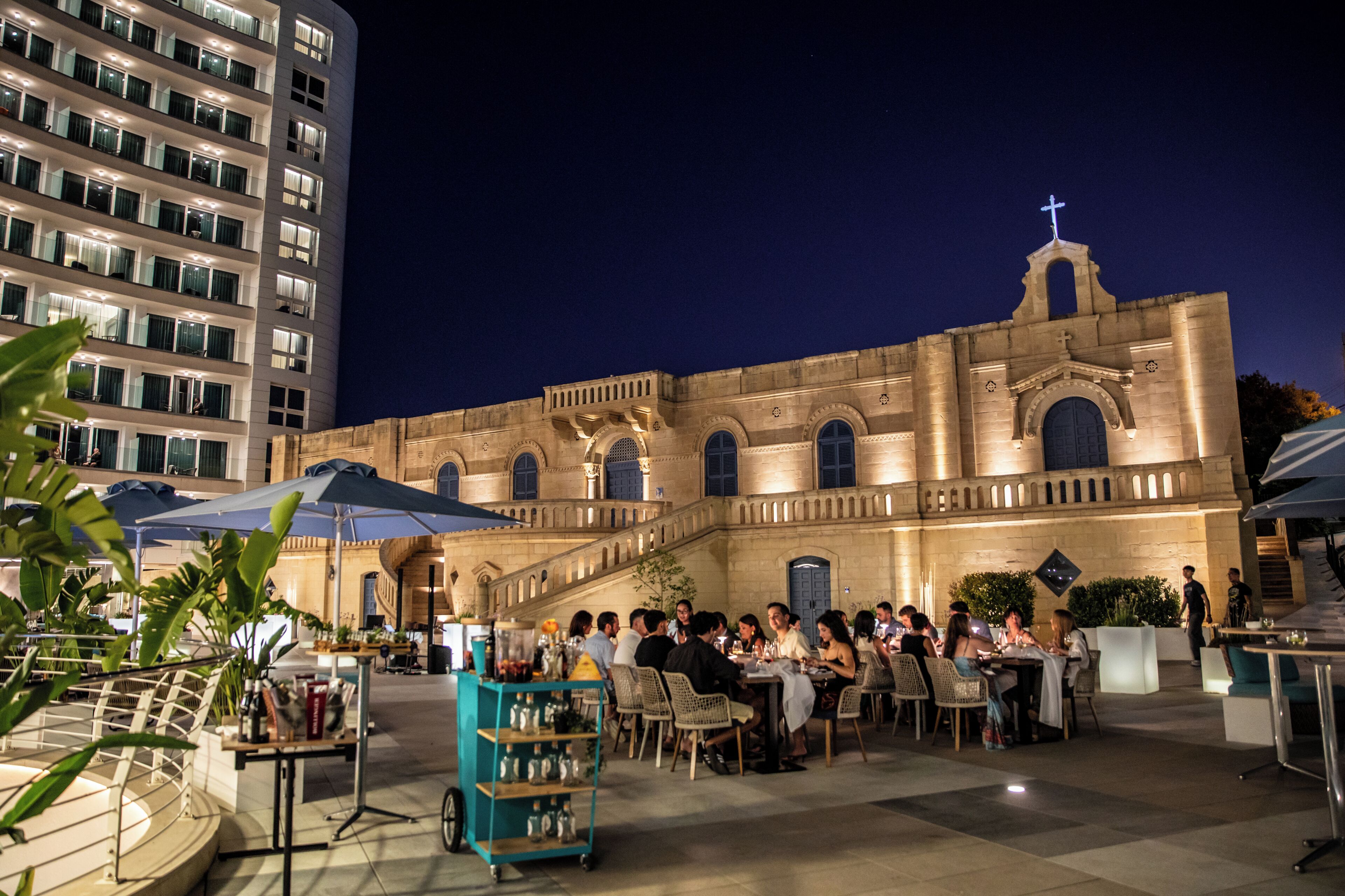 Un área de comedor al aire libre con mesas y sillas, una sombrilla azul y un edificio con ventanas arqueadas y una cruz en la parte superior.