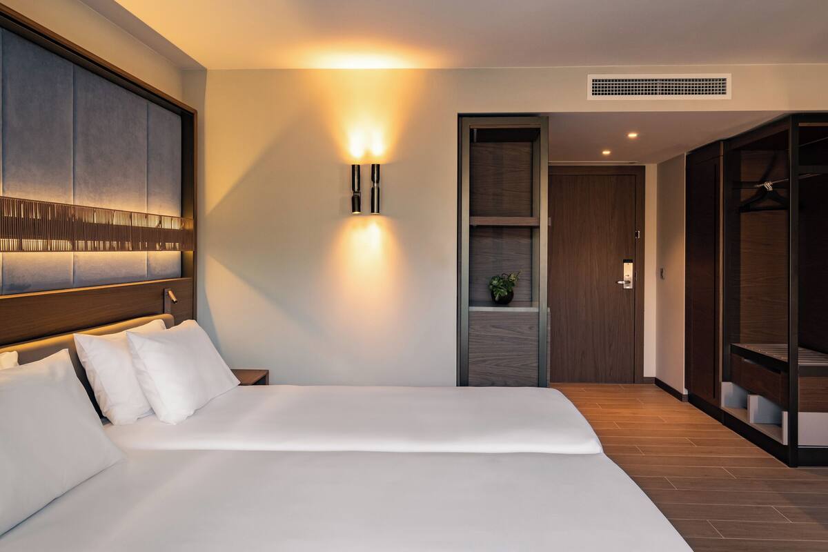 Una habitación de hotel moderna con una cama grande, un aplique de pared, un armario de madera y una planta pequeña.