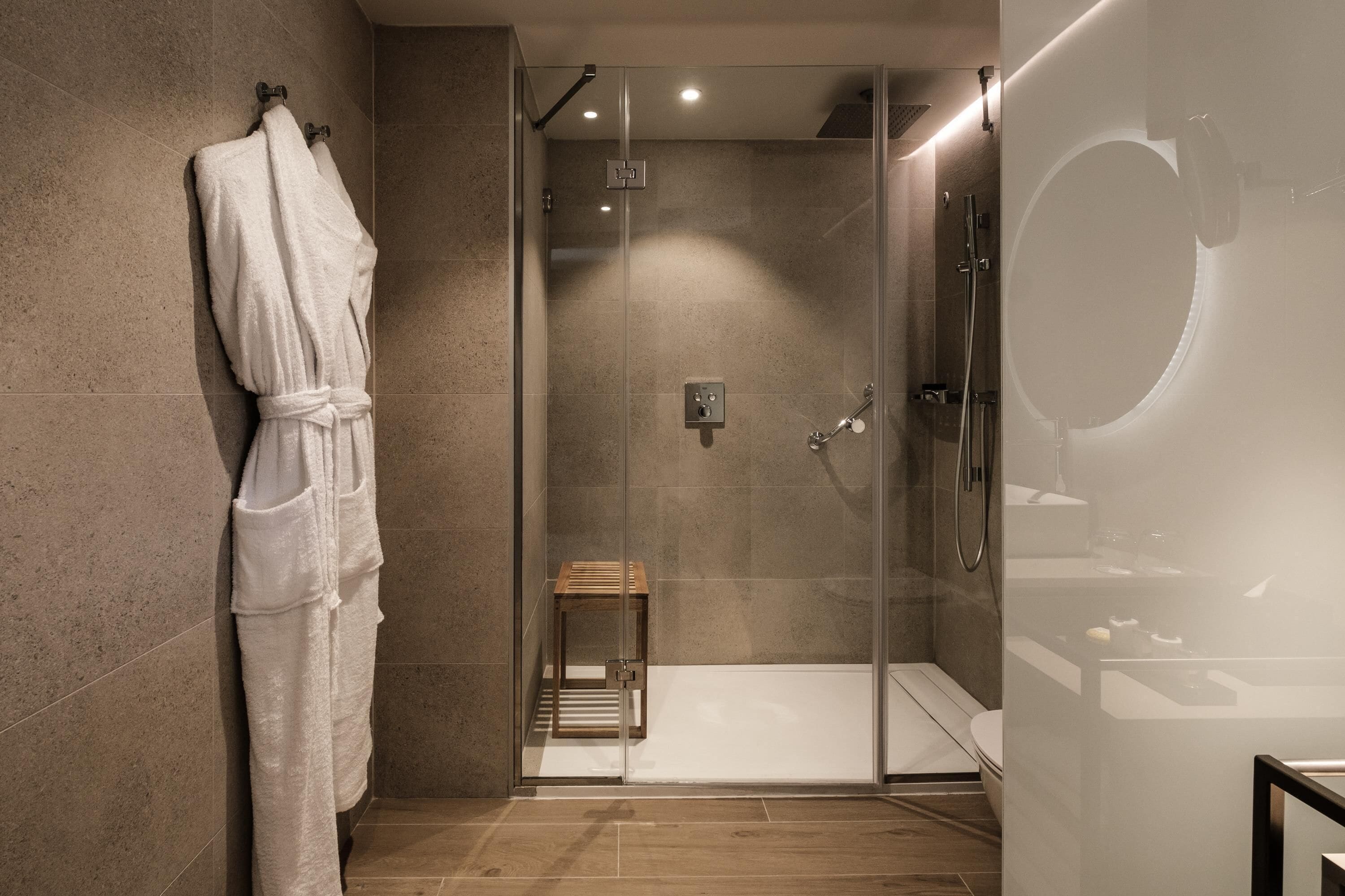 A modern bathroom with a glass shower enclosure, a wooden stool, and a white bathrobe hanging on the wall.