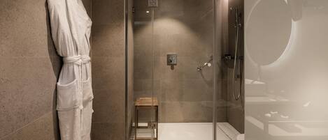 A modern bathroom with a glass shower enclosure, a wooden stool, and a white bathrobe hanging on the wall.