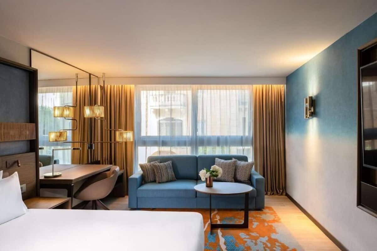 Habitación de hotel con cama, sofá, escritorio y una mesita. Hay cortinas en las ventanas y una lámpara sobre el escritorio.