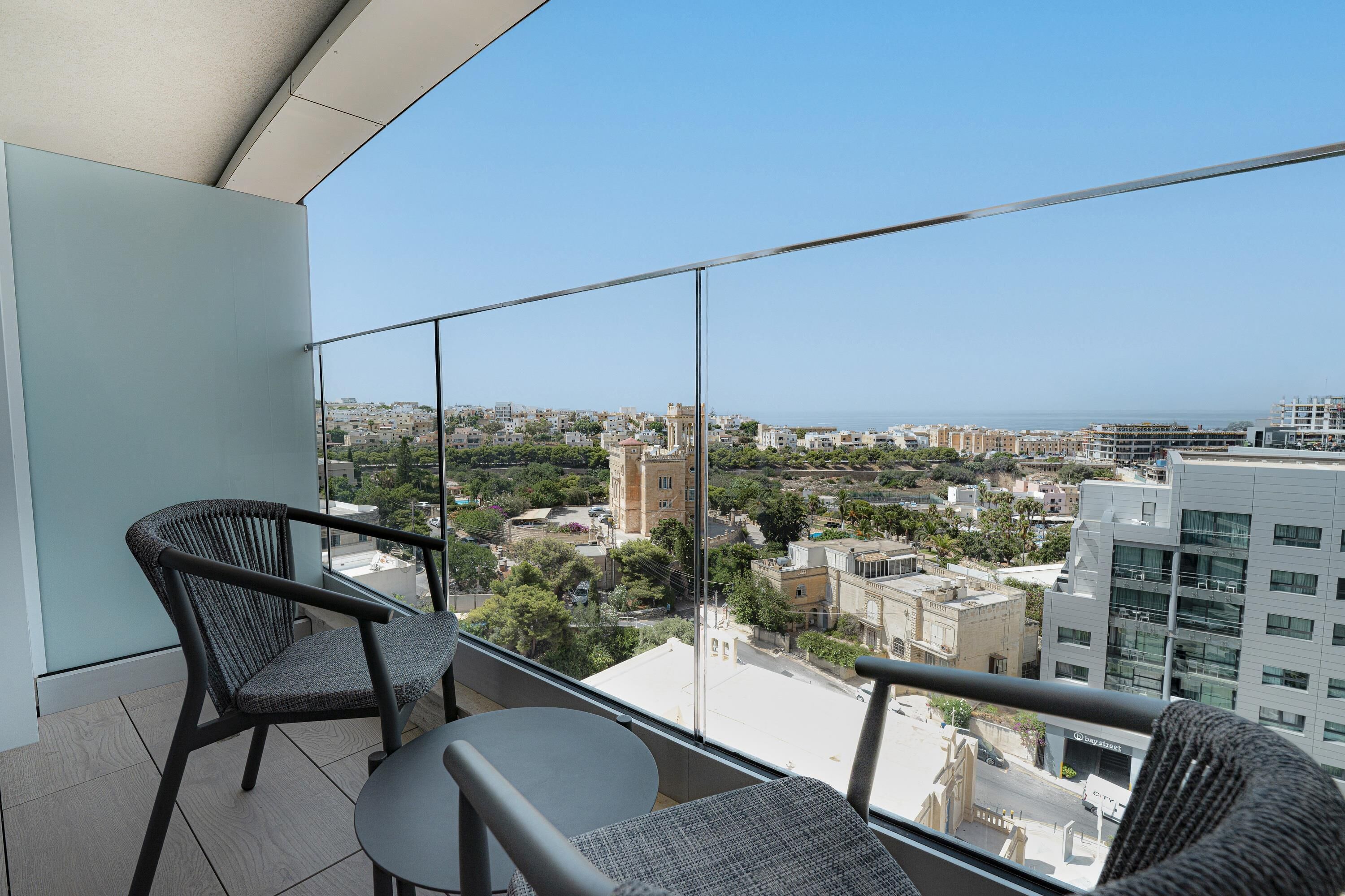 A balcony with two wicker chairs and a small table, offering a view of a cityscape with buildings and a tower.