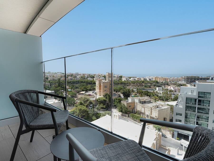 A balcony with two wicker chairs and a small table, offering a view of a cityscape with buildings and a tower.
