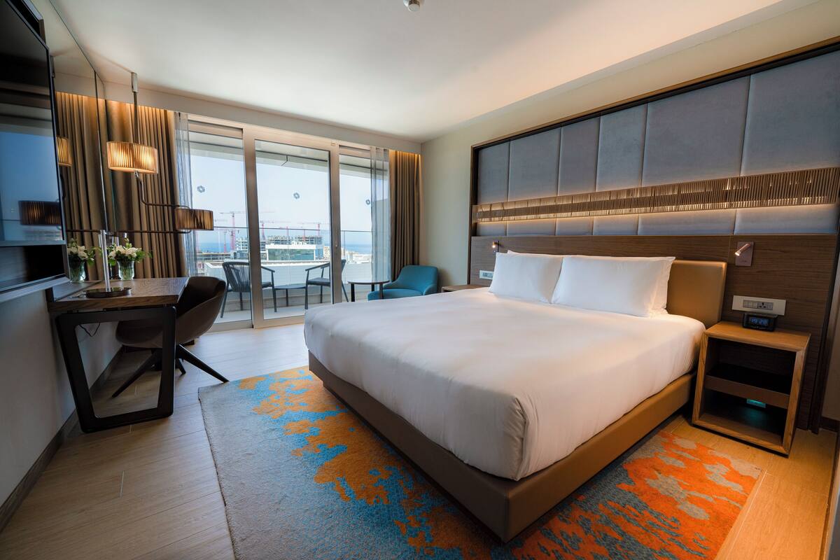 Un hotel moderno con una cama grande, un escritorio, una silla y vistas de la ciudad a través de amplios ventanales.