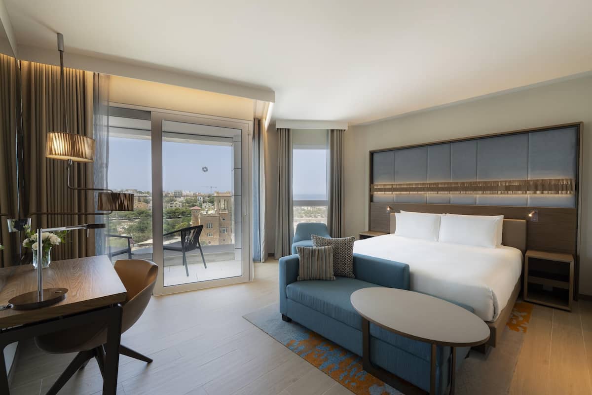 Una habitación de hotel moderna con una cama grande, un sofá, una mesa de comedor y un balcón con vistas a la ciudad.