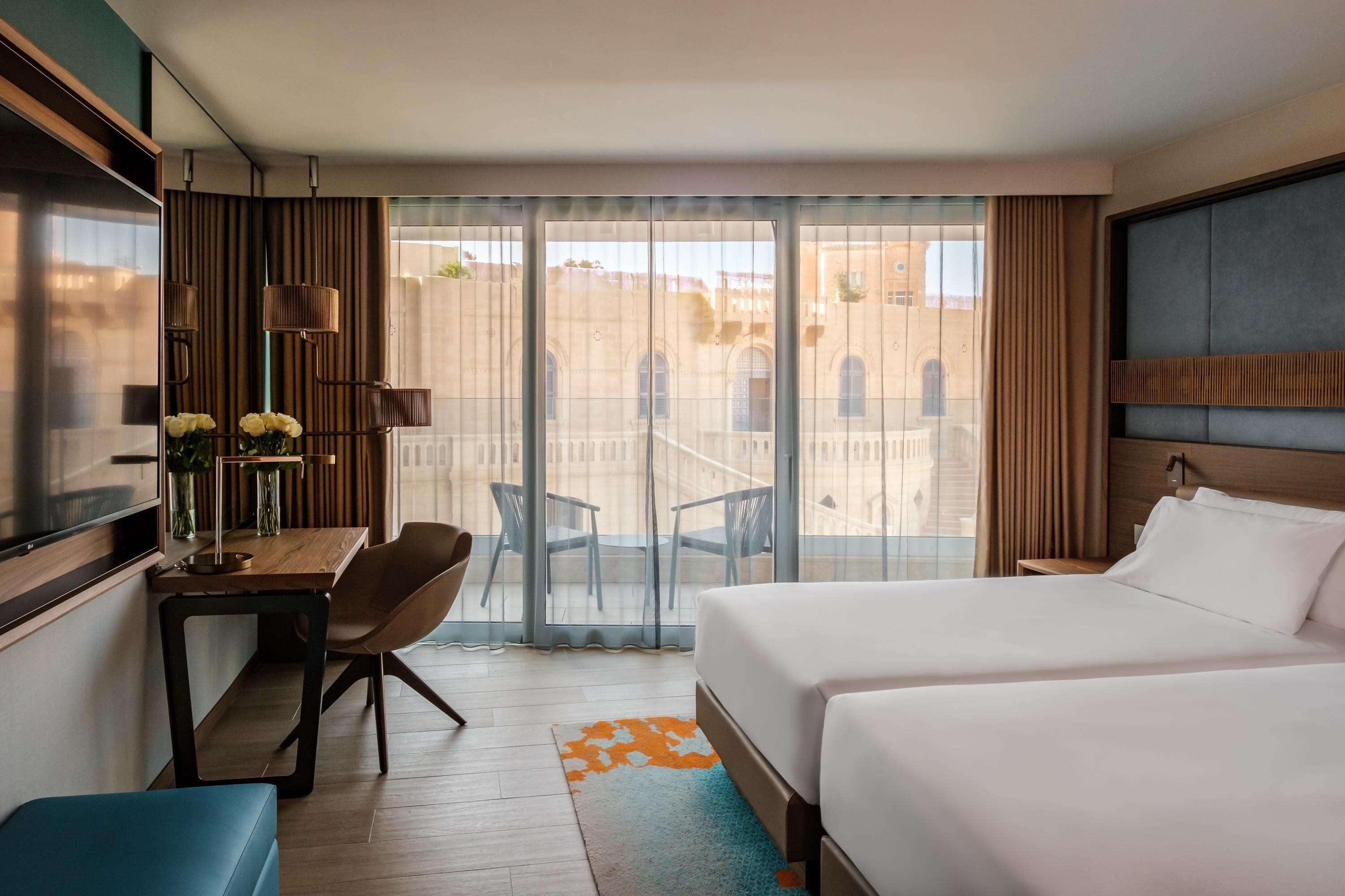 Una habitación de hotel con una cama, un escritorio con silla, un televisor y vistas al exterior a través de amplios ventanales.