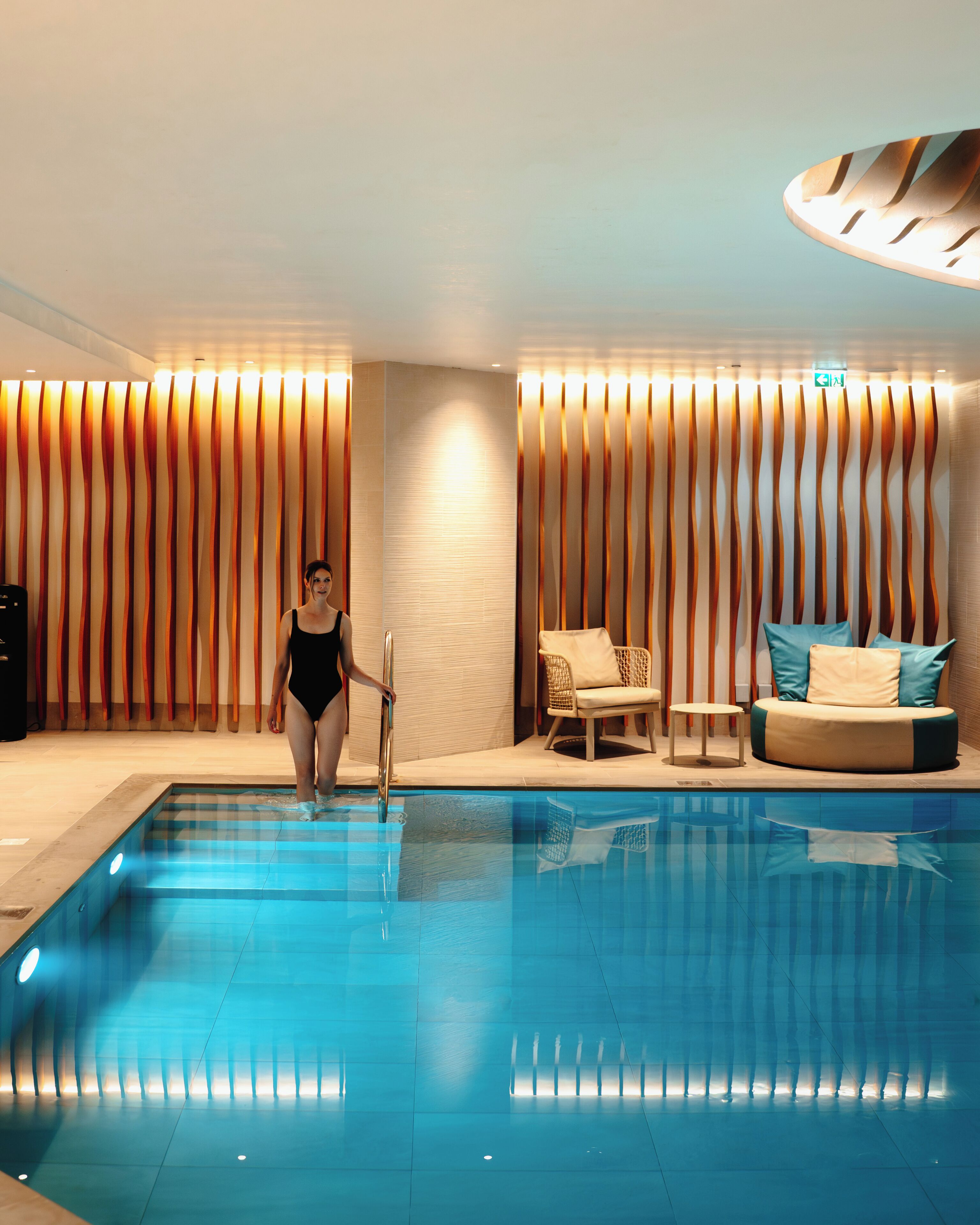 A swimming pool area with a person in a swimsuit, a lounge chair, and a sofa with cushions.