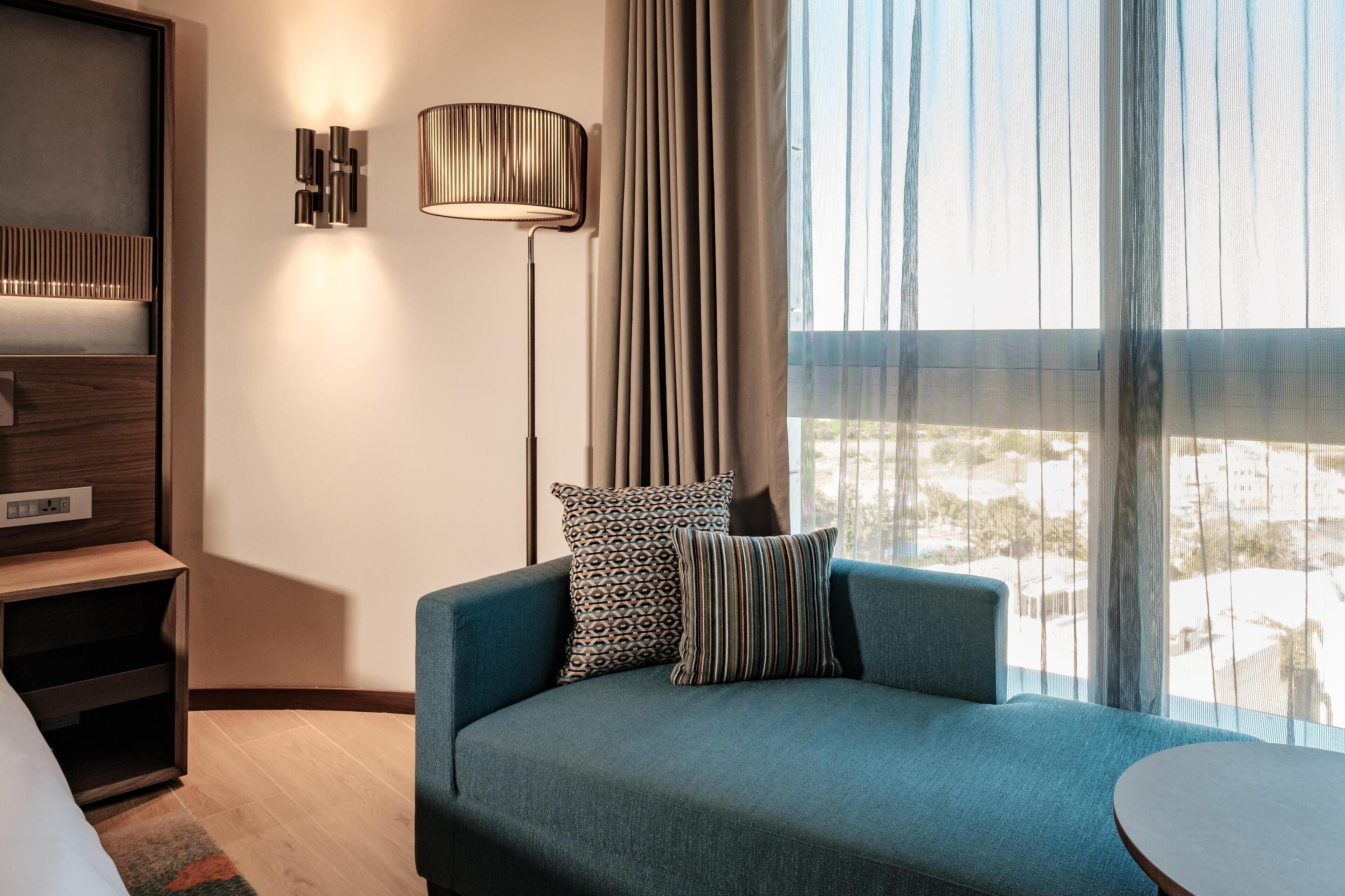 A modern hotel room with a blue sofa, patterned pillows, a standing lamp, and a view of the city through sheer curtains.