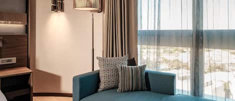 A modern hotel room with a blue sofa, patterned pillows, a standing lamp, and a view of the city through sheer curtains.