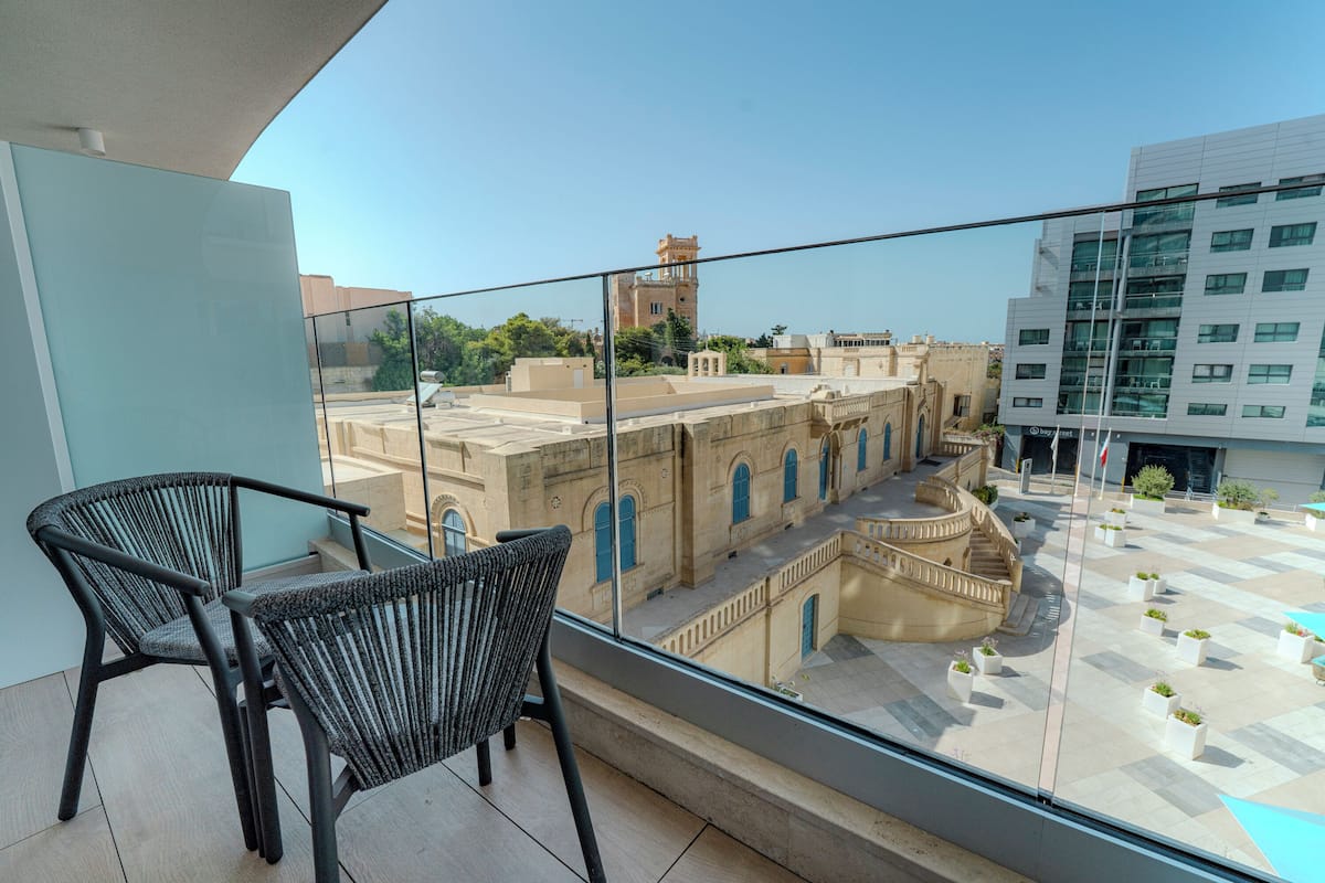 Un balcón con dos sillas de mimbre, vistas a edificios históricos y un edificio moderno con un cartel.