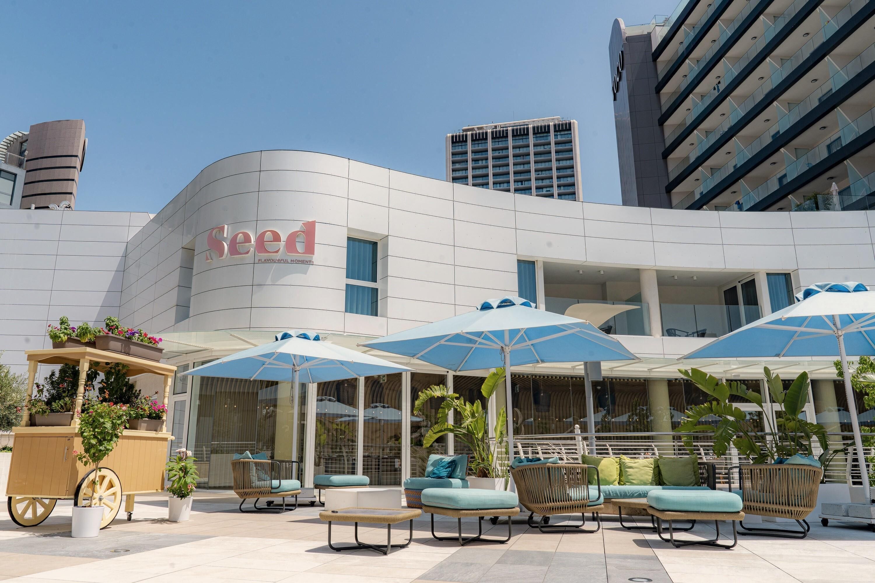 A modern hotel with a curved facade, outdoor seating with umbrellas, and a cart with potted plants.