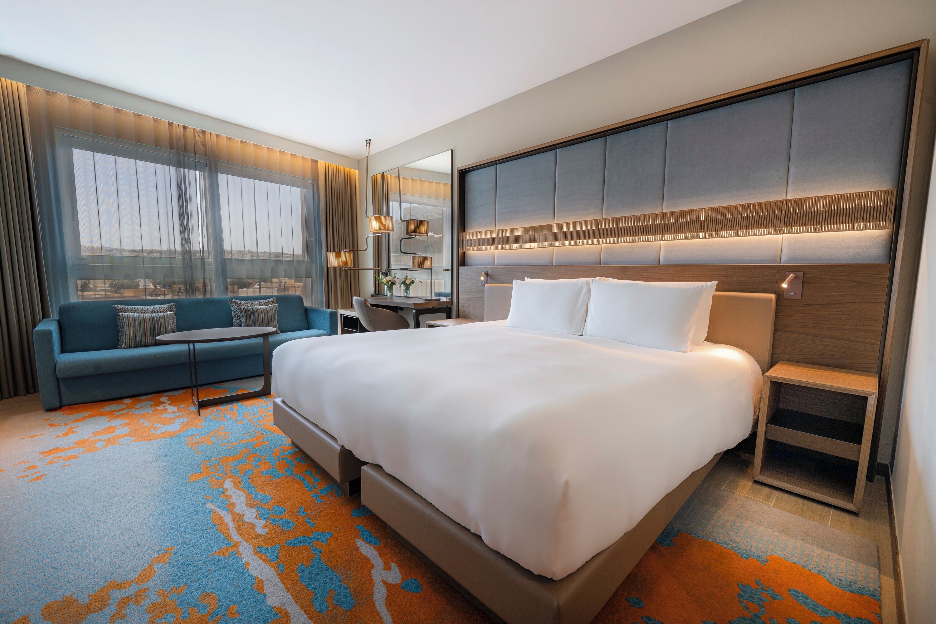 A hotel room with a large bed, a sofa, a desk, and a view of the city.