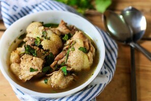 A bowl of chicken and potatoes in a broth, garnished with herbs, on a wooden table with a striped cloth and a spoon. - Exclusive Serene Hotel (Enugu)
