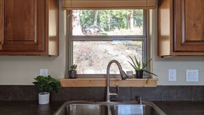 A kitchen with a double stainless steel sink, wooden cabinets, a window with a view of trees, and two potted plants on the windowsill.