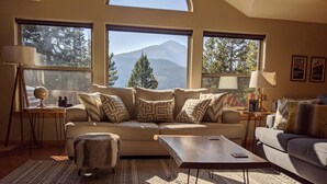 A cozy living room with a sofa, coffee table, and patterned rug. There are two side tables with lamps and a globe. Large windows offer a view of trees and mountains.
