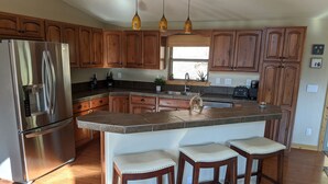 A kitchen with wooden cabinets, a stainless steel refrigerator, a granite island with bar stools, and pendant lighting.