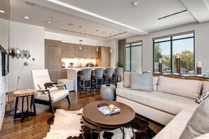 A modern living room with a sofa, coffee table, and chairs. There is a kitchen in the background with bar stools.