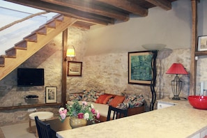 Una habitación acogedora con pared de piedra, escalera de madera, un sofá con cojines estampados, una mesa de comedor con sillas y un tazón rojo sobre la encimera.