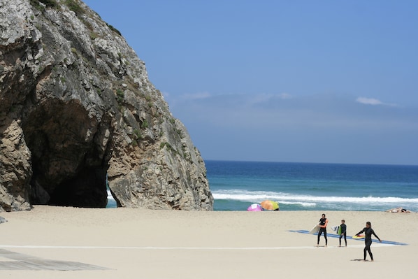 Una playa con una gran formación rocosa, olas del océano y gente con tablas de surf.