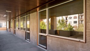 Um corredor com grandes janelas e piso ladrilhado, que leva a um edifício com várias varandas.