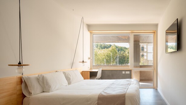 Quarto moderno com uma cama grande, uma mesa de trabalho e vista para um edifício através da janela.