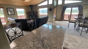 A spacious room with a large marble island, black leather seating, a stone fireplace, and large windows offering views of the outdoors. - The Gateway Cedar Lodge (Alma)
