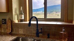 A kitchen with a double sink, a window with a view of a landscape, and a bottle of CARAMELIT on the counter. - The Gateway Cedar Lodge (Alma)
