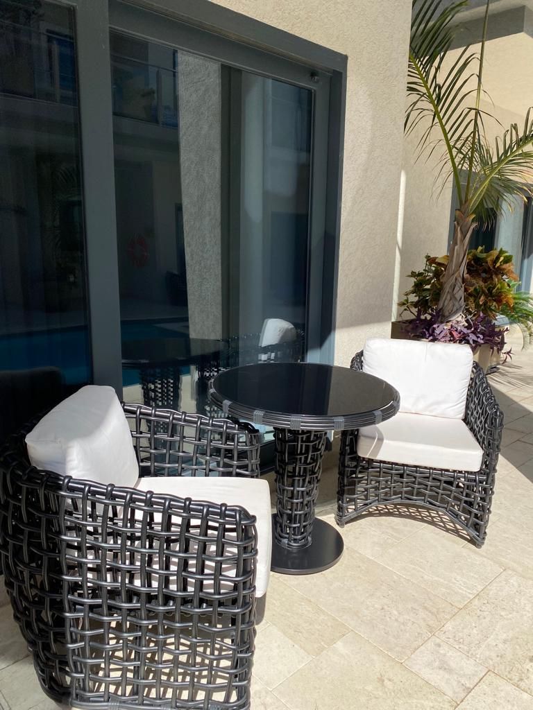 Een terras met rieten meubels en een glazen tafel, naast een gebouw met grote ramen.