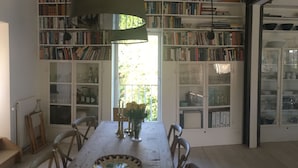 A dining area with a wooden table, chairs, and a hanging light fixture. There are bookshelves filled with books on either side of the room.