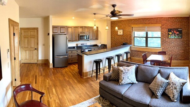 A spacious kitchen with a central island, wooden flooring, and a dining area with a brick wall. - Main Street Retreats 103 Brand New Downtown Apt 103 (Waynesville)