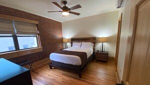 A bedroom with a wooden floor, a bed with white bedding, a ceiling fan, and a window with blinds. - Main Street Retreats 103 Brand New Downtown Apt 103 (Waynesville)