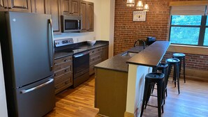 A modern kitchen with wooden cabinets, stainless steel appliances, a central island with stools, and a brick accent wall. - Main Street Retreats 103 Brand New Downtown Apt 103 (Waynesville)